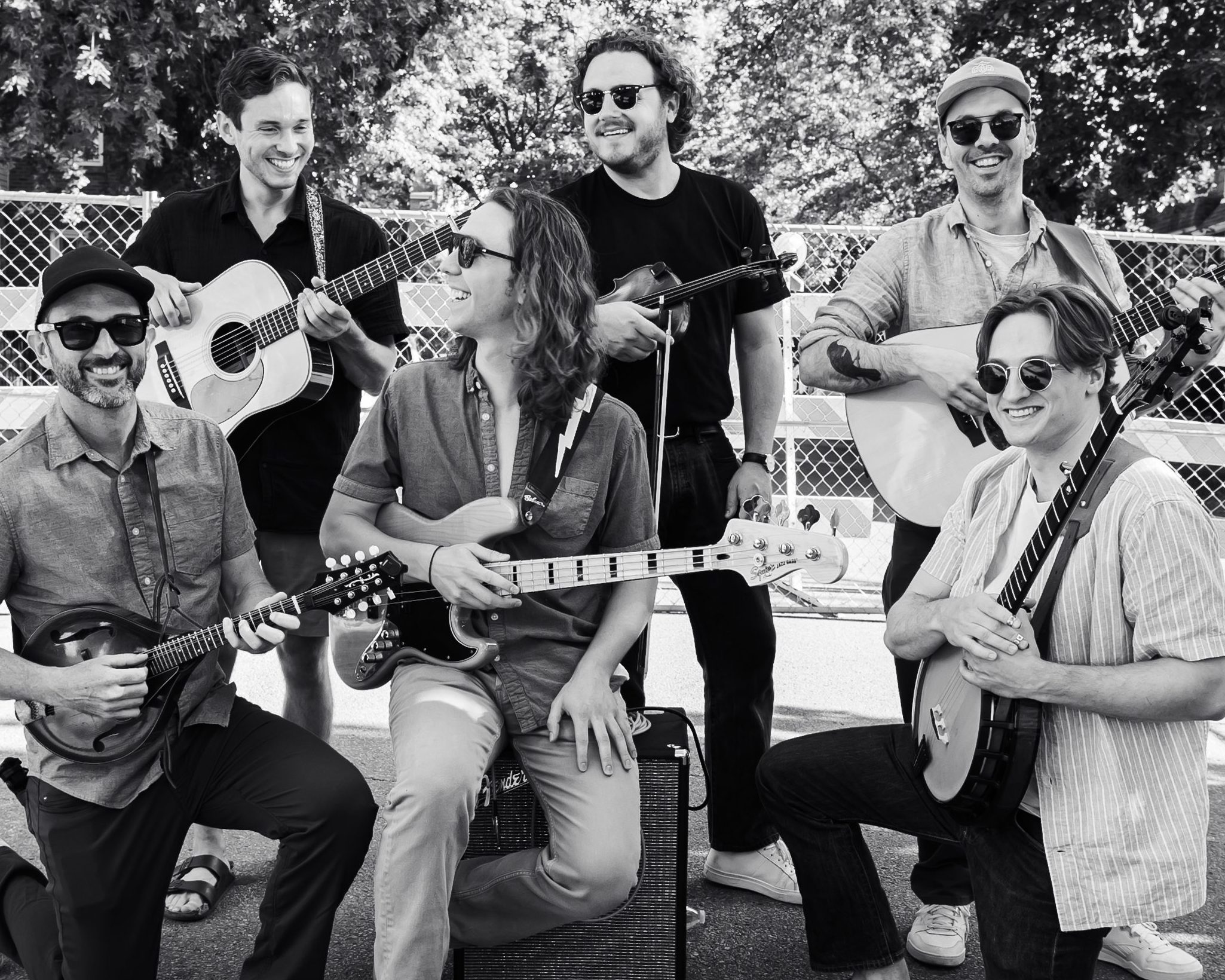 Black and white photo of six male musicians playing instruments outside, smiling at the camera, with medium-length hair an...